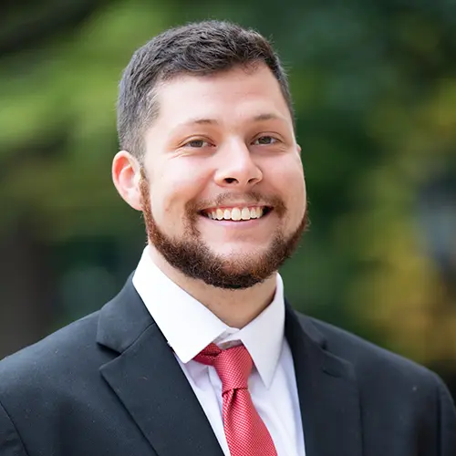 A professional headshot of a man standing in front of an outdoor background with a black blazer and red tie on.