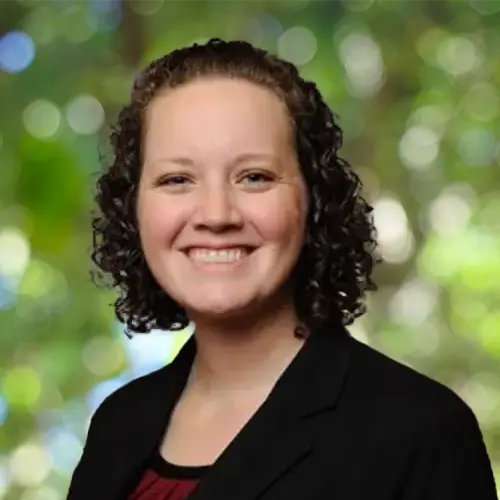 A professional headshot of a brown haired woman smiling in front of a dark backdrop.