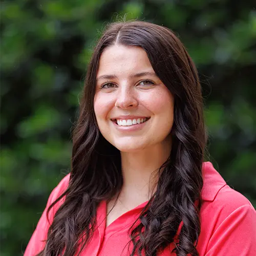 Smiling woman with long dark wavy hair wearing a coral polo shirt, standing in front of a blurred green leafy background.