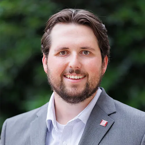 A professional headshot of a man standing in front of an outdoor background with a gray blazer on.