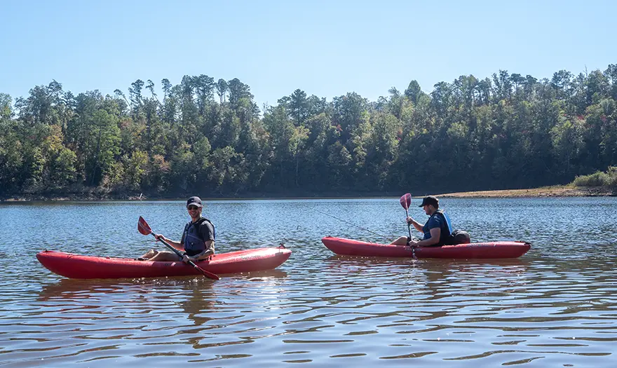 Two people on a kayak paddling down a river with trees and a blue sky in the background.