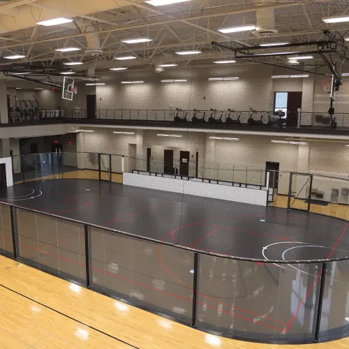 Indoor multipurpose court at a recreation center featuring a black playing surface with red and white lines, surrounded by clear safety panels, with an upper-level track and cardio equipment overlooking the space.